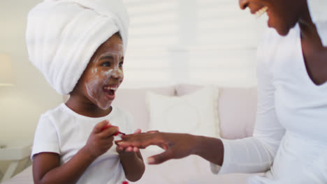 Happy-african-american-mother-and-daughter-sitting-on-bed-and-painting-nails