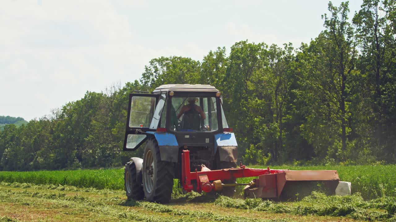 Modern tractor cuts green grass in the countryside. Backside view of an agricultural machine for cutting grass for livestock on the field in a summer day.