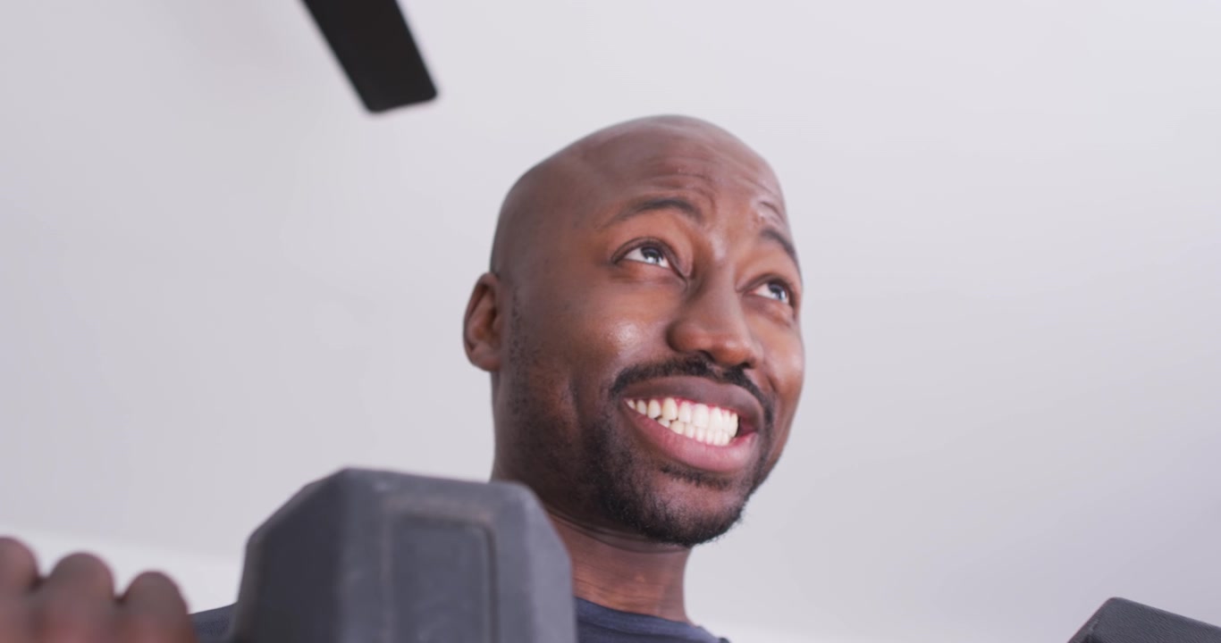 Uomo afroamericano che sorride e poi solleva manubri esagonali a casa durante l'allenamento, ripreso da una telecamera ad angolo basso.