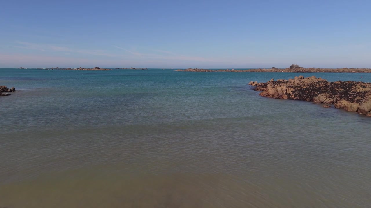 Guernsey, Isole del Canale. Filmato lento e molto basso con drone in avanti su onde leggere che si infrangono a riva, dalla costa a formazioni rocciose, con la bassa marea nel sole del tardo pomeriggio e mare calmo.