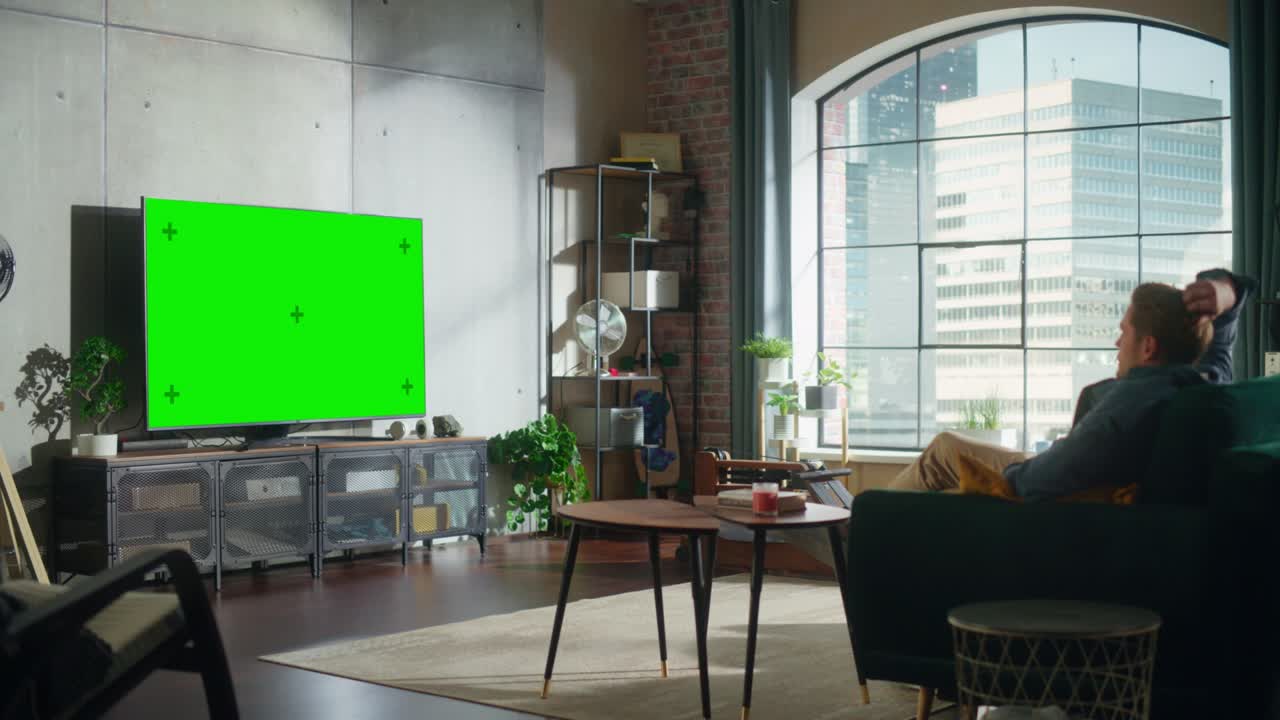 joven hombre guapo sentado en un sofá y viendo la televisión con pantalla verde horizontal mock up. es el tiempo del día en la habitación en casa sala de estar en el apartamento loft.