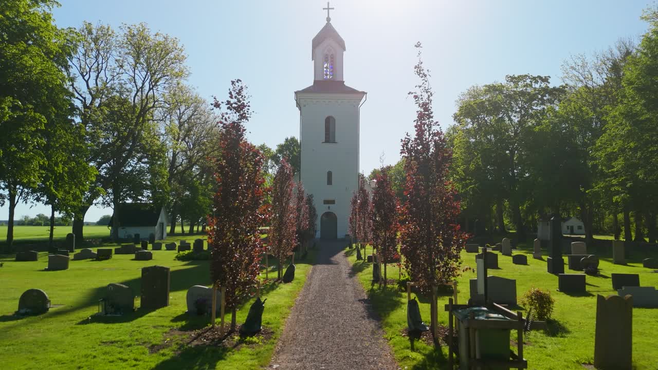 Luchtfoto van een drone die vooruit beweegt langs een met bomen omzoomd kerkhofpad met grafstenen, leidend naar de kerktoren van Grinstad in een zonnig landelijk landschap.