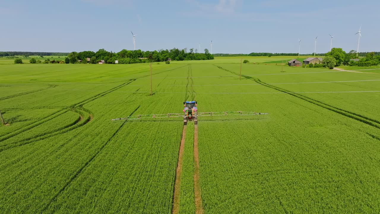 Drone establishing shot shows tractor spraying crop field with turbines, Latvia