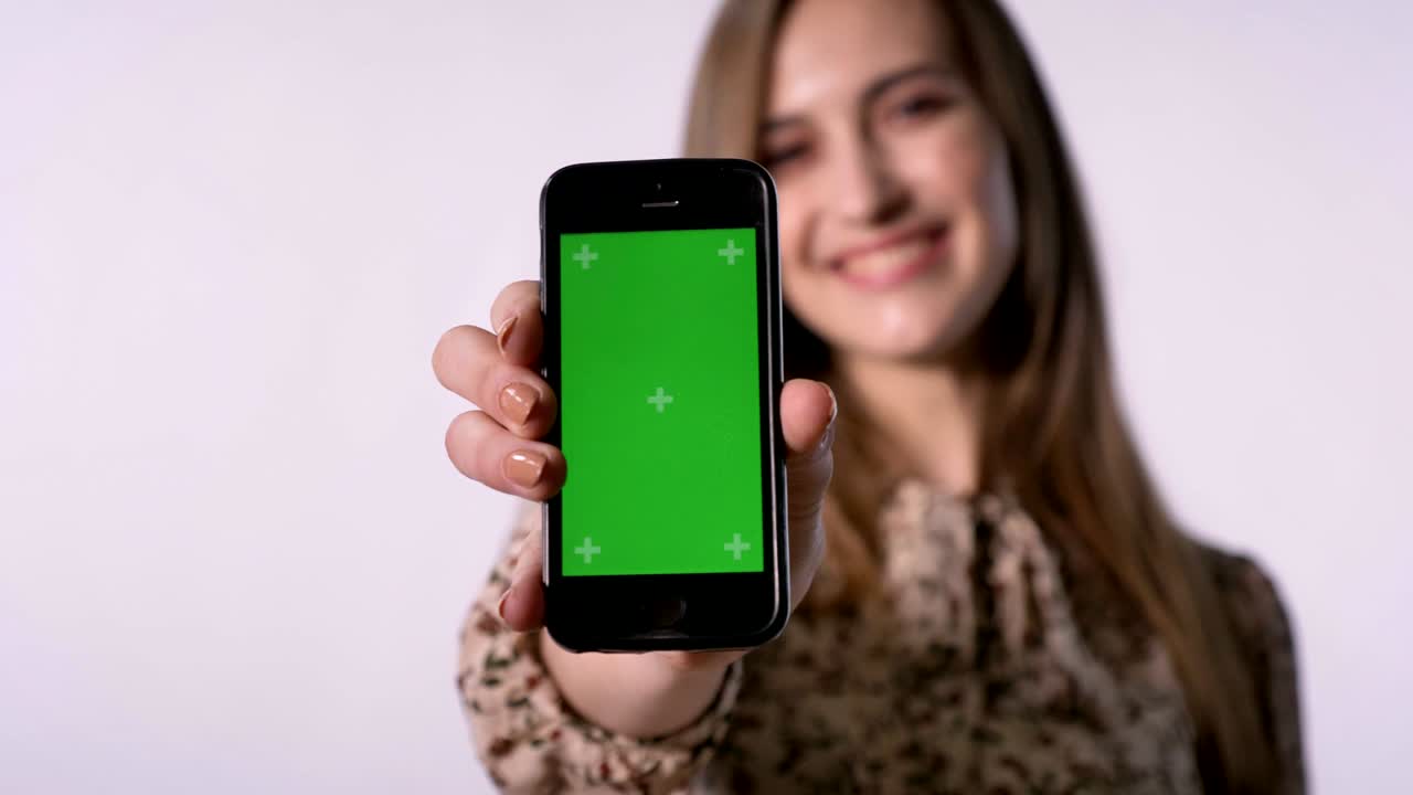 Young cheerful woman shows green screen of smartphone, smiling, watches at camera, communication concept, white background