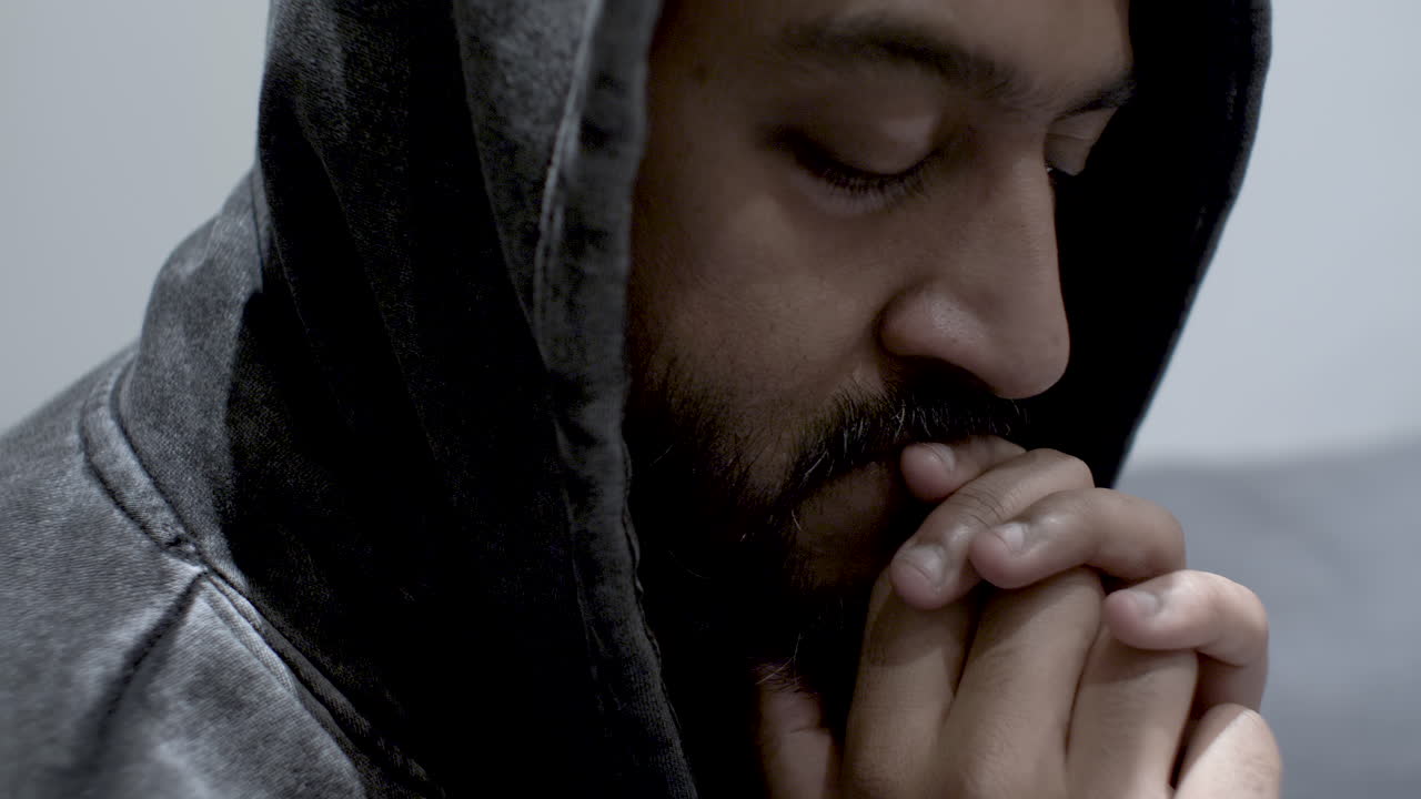 A close up shot of an Ethnic Minority Man holding his hands in front of his mouth, eyes closed as he rocks back and forth wrestling with his deep dark anxious thoughts