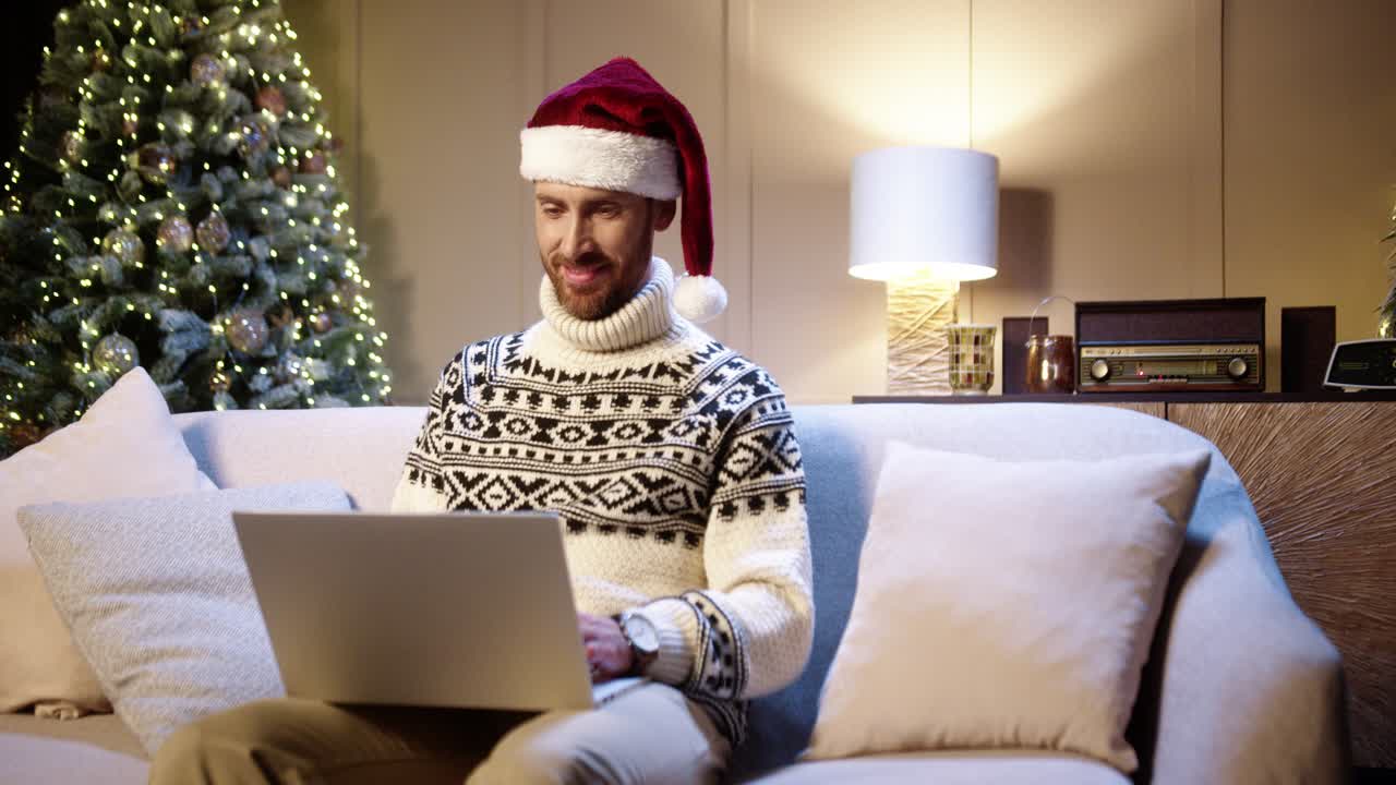 retrato de hombre feliz y guapo con sombrero de santa sentado en una habitación acogedora con árbol de navidad y escribiendo en una laptop mirando hacia otro lado