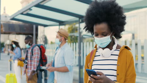 mujer afroamericana con máscara facial y auriculares usando un teléfono inteligente en la parada de autobús mientras otros viajeros esperan el transporte