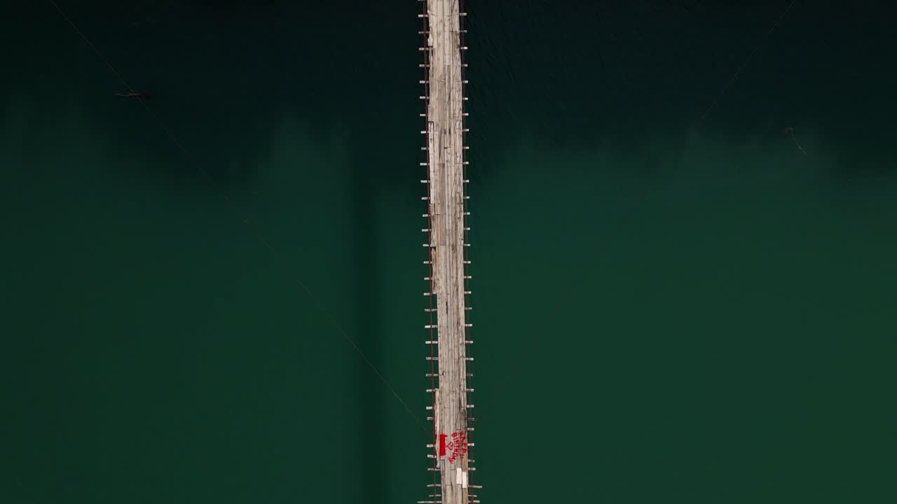 Aerial drone view of a suspension bridge over turquoise water surrounded by green mountains in Albania. Scenic and adventurous summer landscape