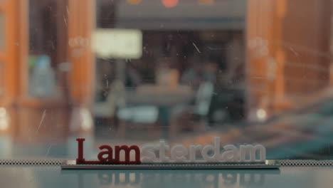 Blick-Auf-Die-Kleine-Plastikfigur-Der-Iamsterdam-Buchstabenskulptur-Auf-Der-Brücke-Vor-Verschwommenem-Stadtbild-In-Amsterdam,-Niederlande