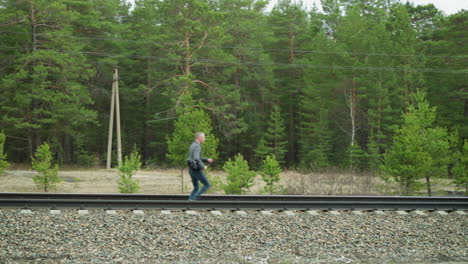 a man in a grey suit jacket and blue jeans is seen running slowly along railway tracks, surrounded by lush green forest and yellow poles