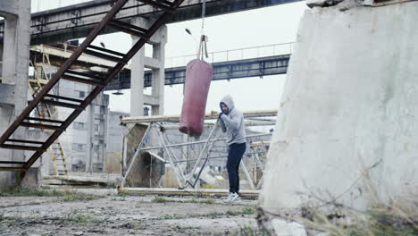 fernsicht auf einen kaukasischen mann in grauem hoodie, der an einem bewölkten morgen einen boxsack im freien einer verlassenen fabrik schlägt