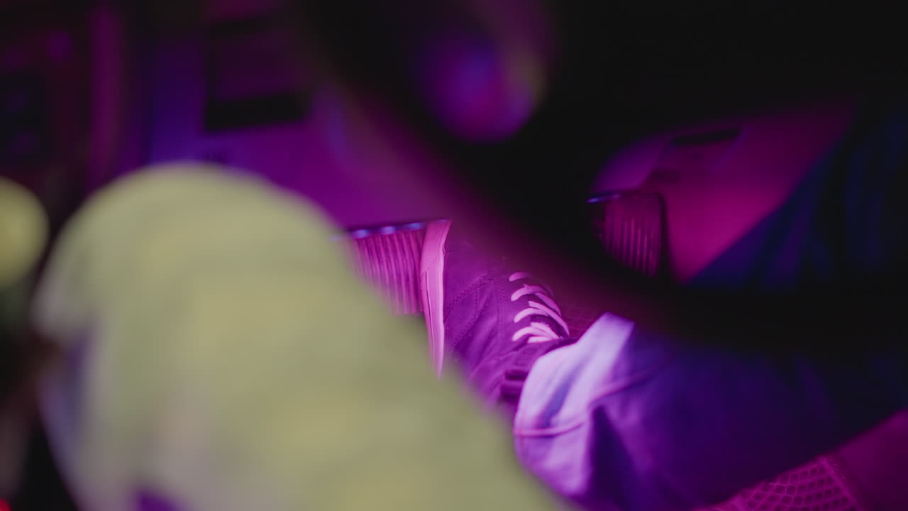 primo piano notturno di sneaker bianche nel vano piedi del passeggero, tenue illuminazione viola al neon su risvolto in denim e suola della scarpa, intimità sul sedile posteriore, texture del rivestimento del sedile, atmosfera con bassi leggeri, fotogrammi che ritraggono un frequentatore di club a riposo