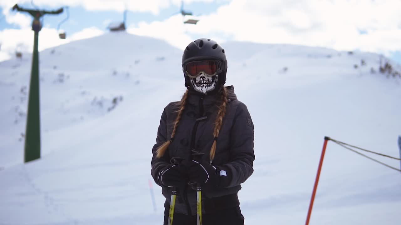 Young woman with long braids and skeleton mask wearing black ski suit on a ski slope in front of ski lift