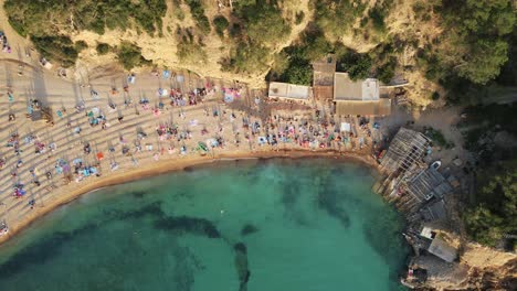 cinematic aerial shot of tourists relaxing and sunbathing at the turquoise seashore of cala benirras in spanish island of ibiza