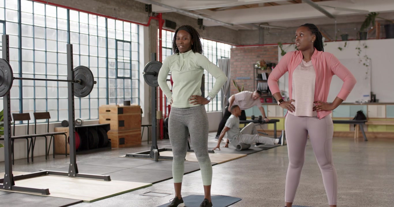 Female workout partners standing on mats near squat rack, waiting for coach cue and preparing