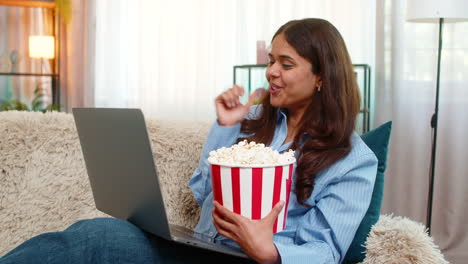 indian woman watching movie with popcorn smiling at laptop screen on home sofa during cozy moment