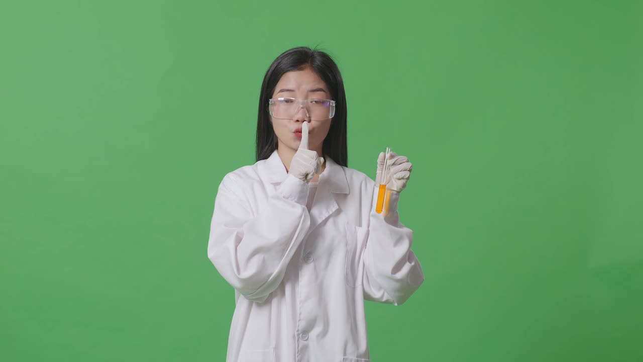 실험관 안의 오렌지색 액체와 함께 카메라를 바라보고 실험실의 녹색 스크린 배경에 서있는 동안 shh 제스처를 만드는 아시아 여성 과학자