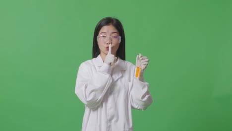 asian woman scientist with orange liquid in the test tube looking at camera and making shh gesture while standing on the green screen background in the laboratory