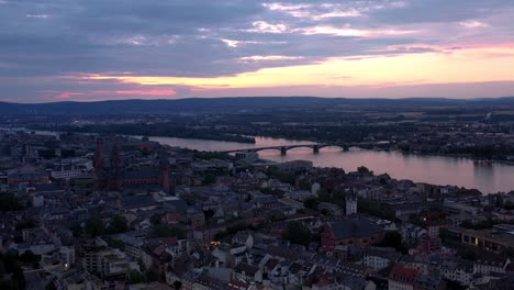 Seitliche-Drohnenaufnahme-Von-Mainz-Zur-Magischen-Stunde-Nacht,-Die-Um-Das-Stadtzentrum-Kreist,-Mit-Dem-Dom-Und-Dem-Dunklen-Rheinwasser-Im-Hintergrund,-Das-Einen-Farbenfrohen-Himmel-Zeigt