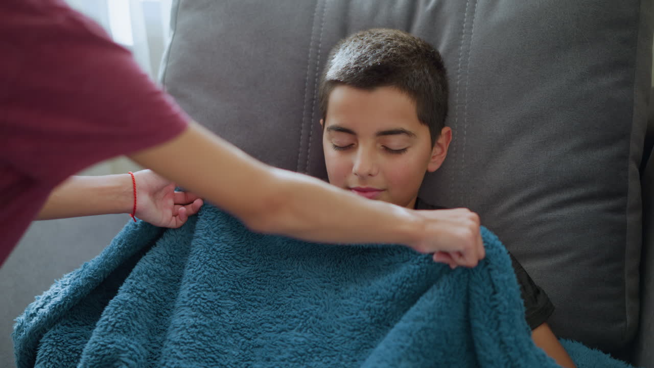 Close-up of cute boy sleeping on couch, smiling peacefully, being covered with a fluffy blanket by someone, with soft natural light filtering through the window