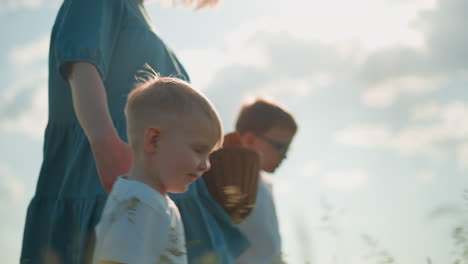 a tender moment in a grass field, focusing on a young boy smiling while walking with his mother in a blue gown and another child, both in white shirts. the woman face is not clearly visible