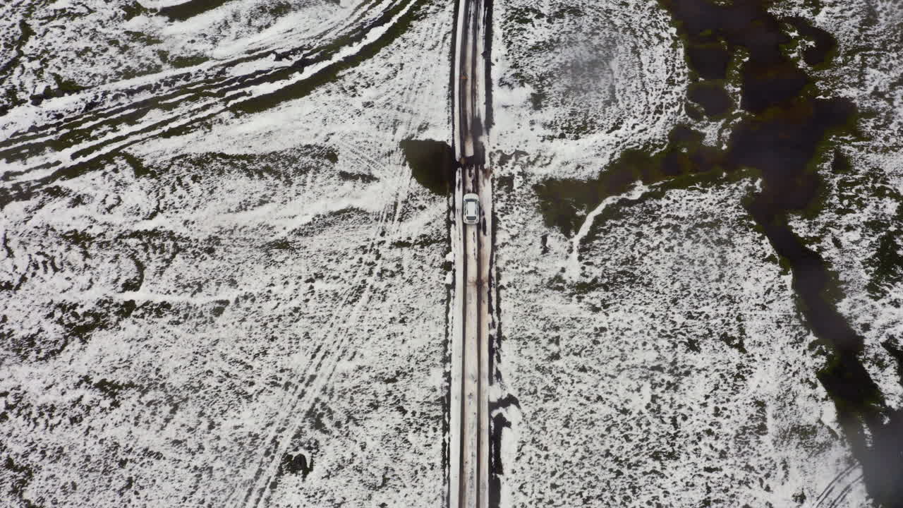Ripresa aerea dall'alto a seguire di un'auto che percorre una strada di montagna in Islanda, parzialmente coperta di neve.