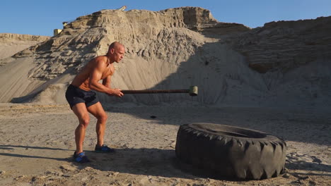 bald man strongman hits a hammer on a huge wheel in the sandy mountains in slow motion. strength and endurance training for wrestlers.