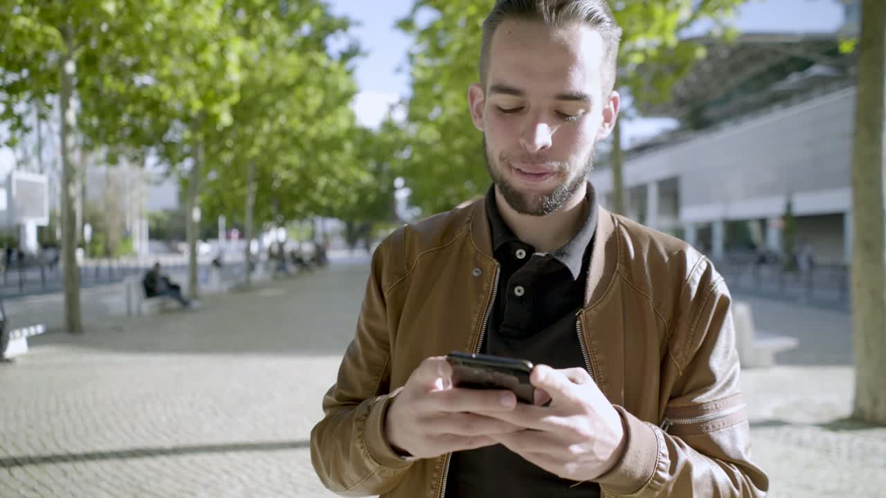 Smiling young bearded man using smartphone during stroll.