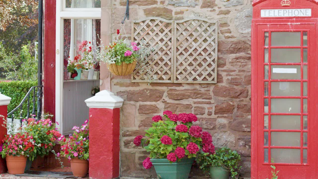 Classic red telephone box beside stone house with vibrant flowers and lush garden in daylight