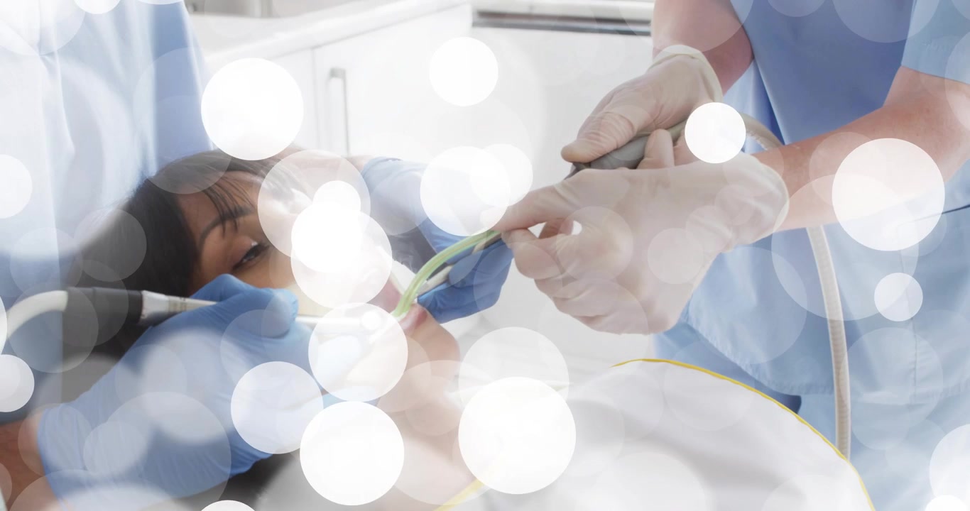 Paciente reclinado sob a lâmpada odontológica, dentista usando espelho e assistente sugando tártaro.