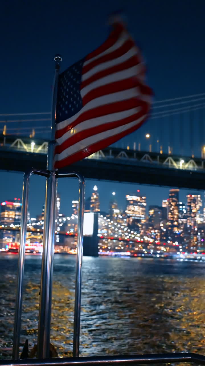 Bandiera americana che sventola al vento a prua della barca. Avvicinandosi al Ponte di Manhattan dal fiume di notte. Video verticale.
