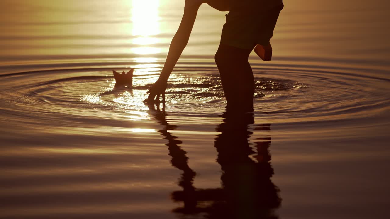Boy launches Red and green Paper Ships in the river. Origami Paper Boats in kid's hands. Beautiful Waves Ripple Background. Travel and nature Concept. Video at sunset.