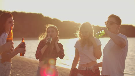 five teenagers celebrate a birthday on the beach party with beer and good mood. they drink beer and dance in the hot evening. this is carefree party at sunset.