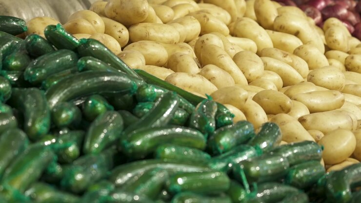 Neatly stacked potatoes and plastic-wrapped cucumbers fill a produce display inside a bright supermarket food store.