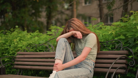 young woman in jeans and white sneakers sits on wooden bench in park, resting her head on her arm in a reflective, somber posture, surrounded by lush greenery and trees, she appears deep in thought