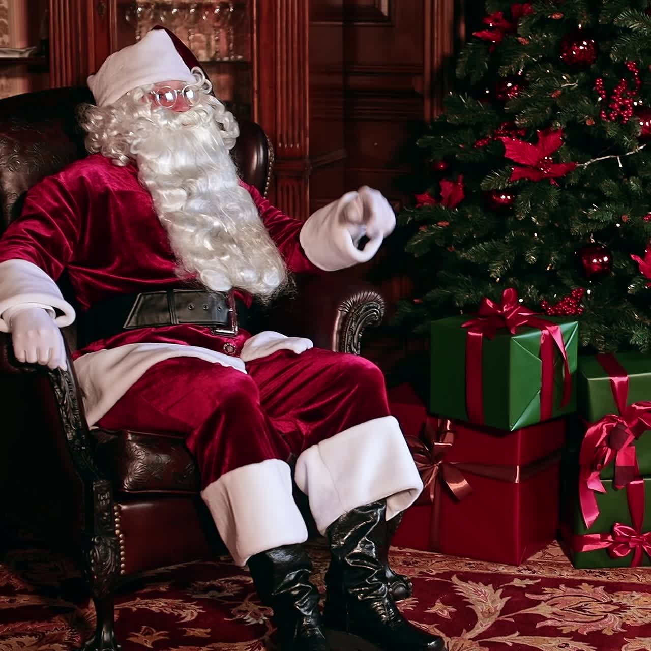 Pensive Santa Claus sitting in a comfortable chair near the Christmas tree with lots of presents around. Holidays preparation concept