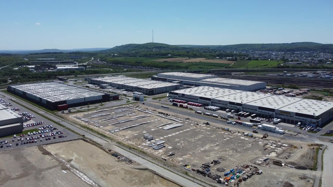 Riprese con drone di una zona industriale con magazzini e un cantiere, circondata da uno scalo ferroviario, un villaggio e piccole colline sotto un cielo soleggiato.