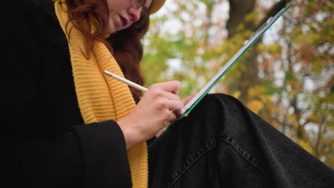 un primer plano de una joven con gafas de sol, boina amarilla y pañuelo amarillo profundamente enfocada en su dibujo, capturando la concentración artística y la creatividad