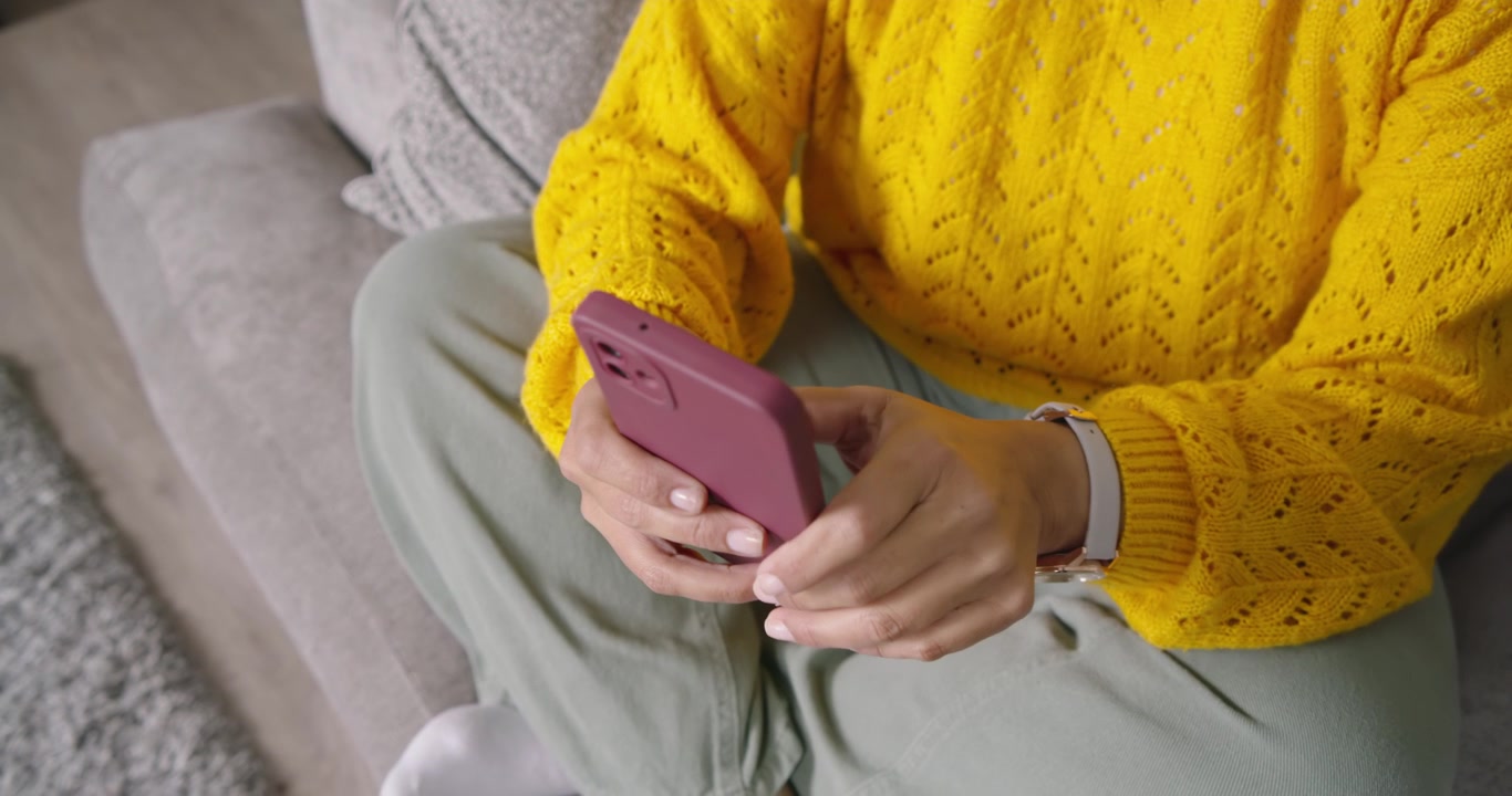 Donna africana matura seduta sul divano con un maglione giallo, che tiene in mano un telefono color malva e controlla i messaggi.