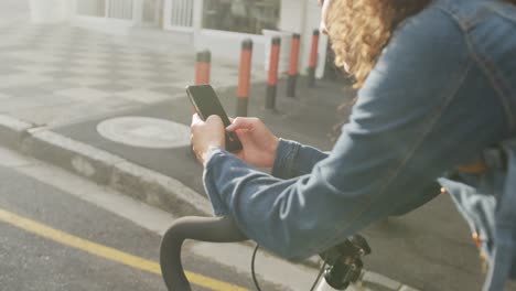 happy biracial woman in city, sitting on bike using smartphone