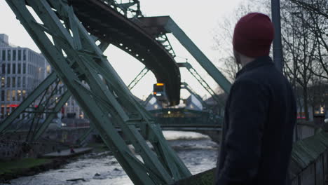 a man observing wuppertal's suspended train pass by in germany at dusk