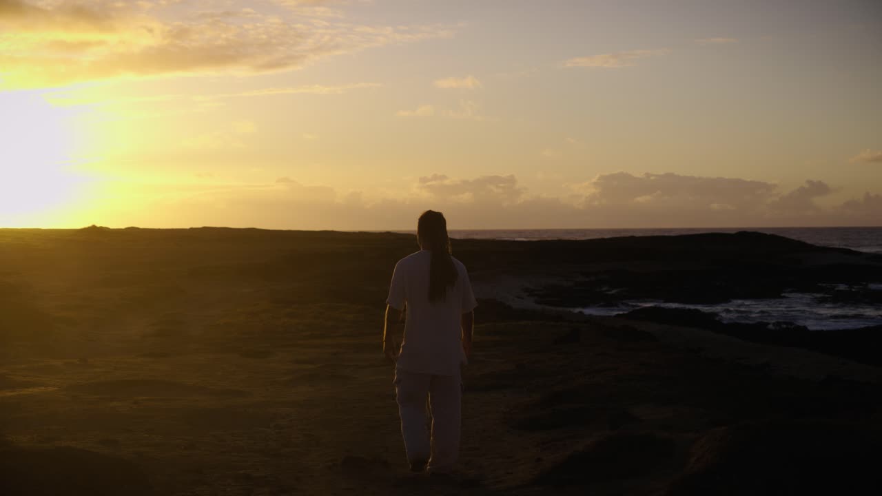 A man strides forward along a rugged coastal path, backlit by the golden sunrise as waves crash in the distance and the horizon glows warmly