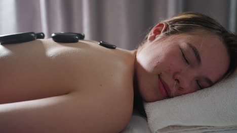 close view of client smiling peacefully with eyes closed while lying on towel with hot black massage stones placed along back during calming therapy in softly lit tranquil spa room near gray curtains