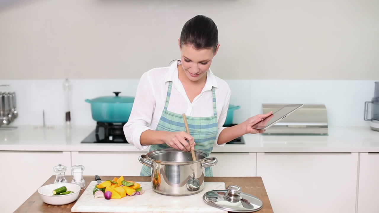 hermosa morena siguiendo una receta en su tableta mientras hace la cena