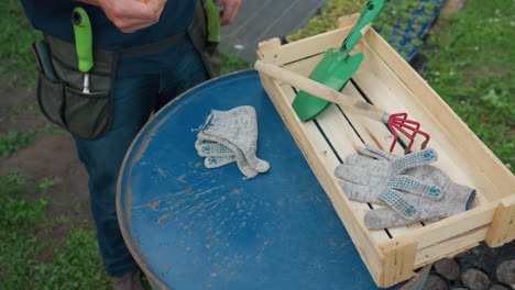 top down view of gardener standing beside blue barrel organizing tools into waist apron pocket with gloves, shovel, hand rake, cutter, and wooden crate visible