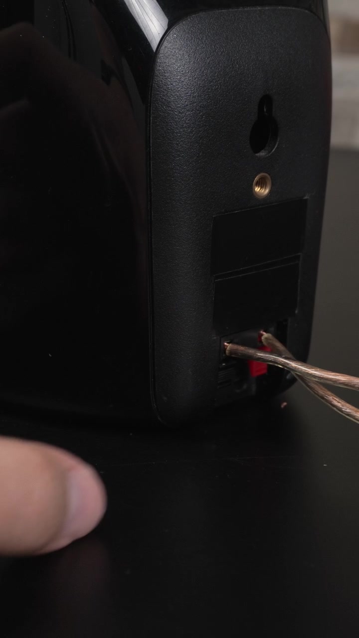 Hands attach bare speaker wire to the spring clip terminals on the back of a black bookshelf speaker on a desk, useful for tutorials, product demos, or home o guides.
