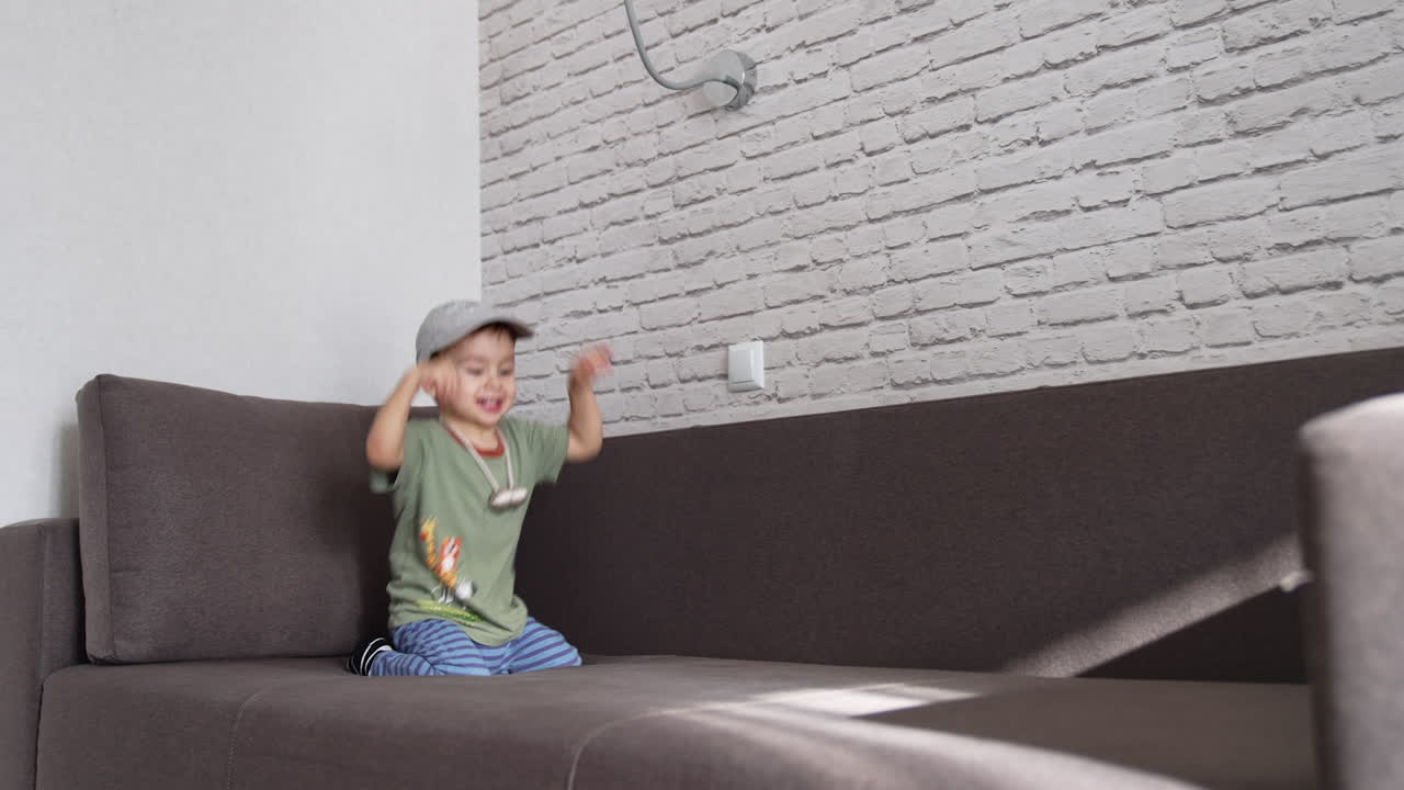 Cute smiley kid in cap sitting on the sofa. Baby boy jumps happily on the sofa at home.