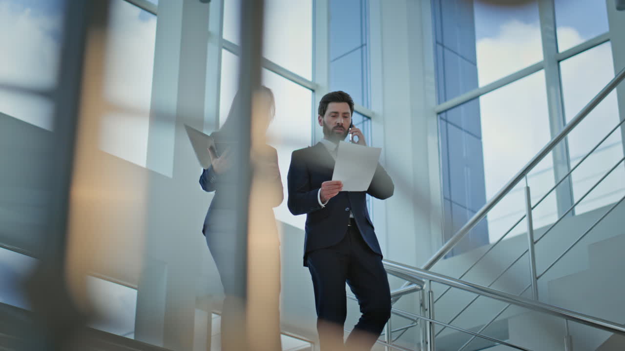 Two business professionals walking down office staircase. Man talking smartphone