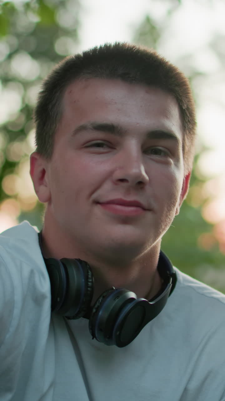 목에 검은 헤드폰을 달고 색 을 입고 야외에 앉아있는 남자의 클로즈업, 얼굴에 미묘한 미소, 녹색 나무와 부드러운 야외 조명으로 둘러싸여