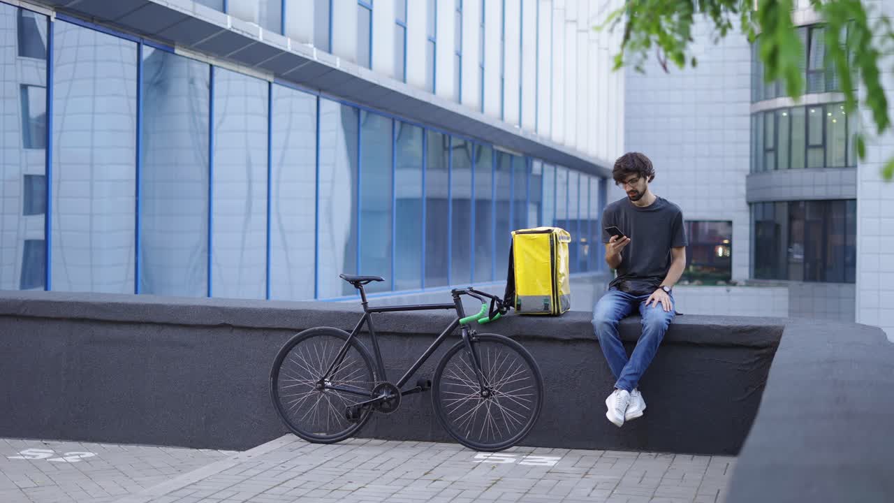 Delivery man have a rest near skyscraper, uses smartphone